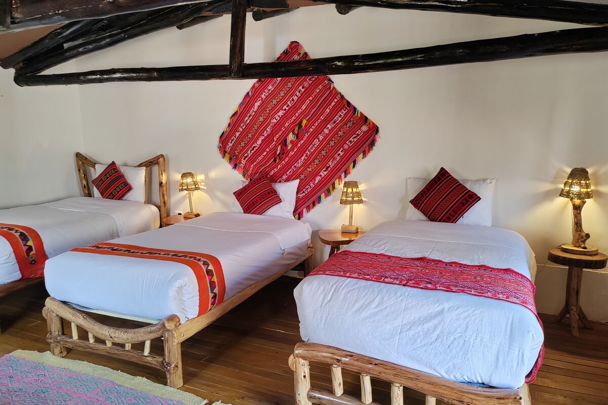 Una habitación con dos camas de madera, cada una con un colchón blanco y colchas rojas estampadas. Hay dos mesitas de noche con lámparas, y un tapiz rojo estampado sobre las camas.
