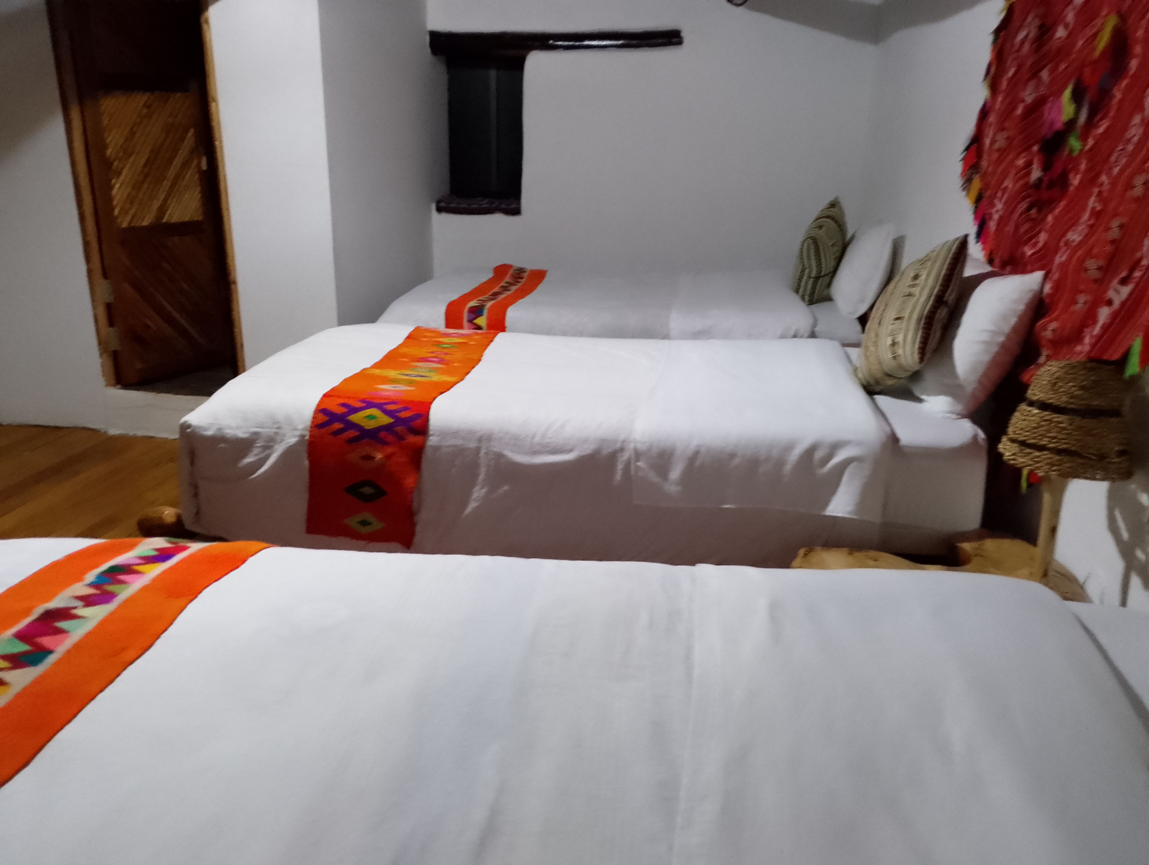 Une chambre avec deux lits, chacun recouvert d’un couvre-lit blanc et d’une couverture colorée. On y trouve une porte en bois et une petite fenêtre avec un rideau.