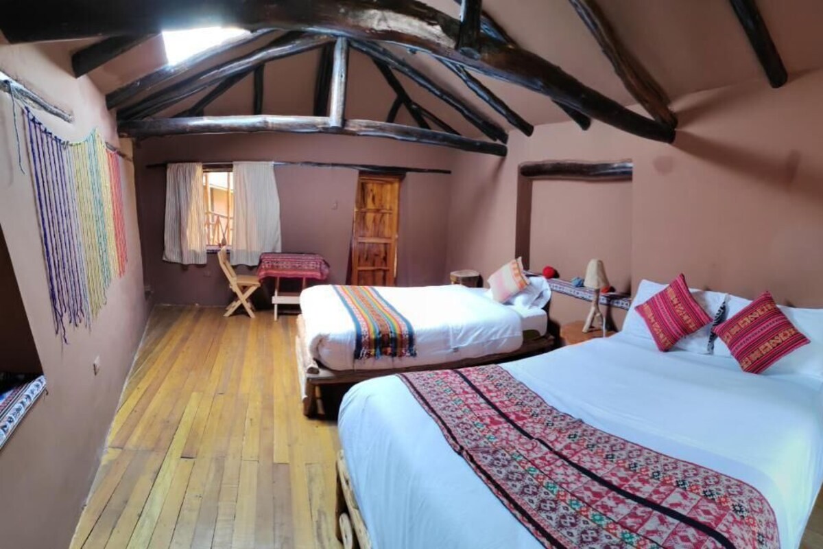 Una habitación acogedora con piso de madera, una cama con ropa de cama estampada, una mesita con mantel rojo y una silla.