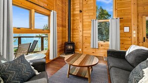 Ein gemütliches Blockhaus-Interieur mit Kamin, großen Fenstern und einer Terrasse mit Blick auf ein Gewässer.