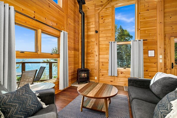 A cozy cabin interior with a fireplace, large windows, and a deck overlooking a body of water.