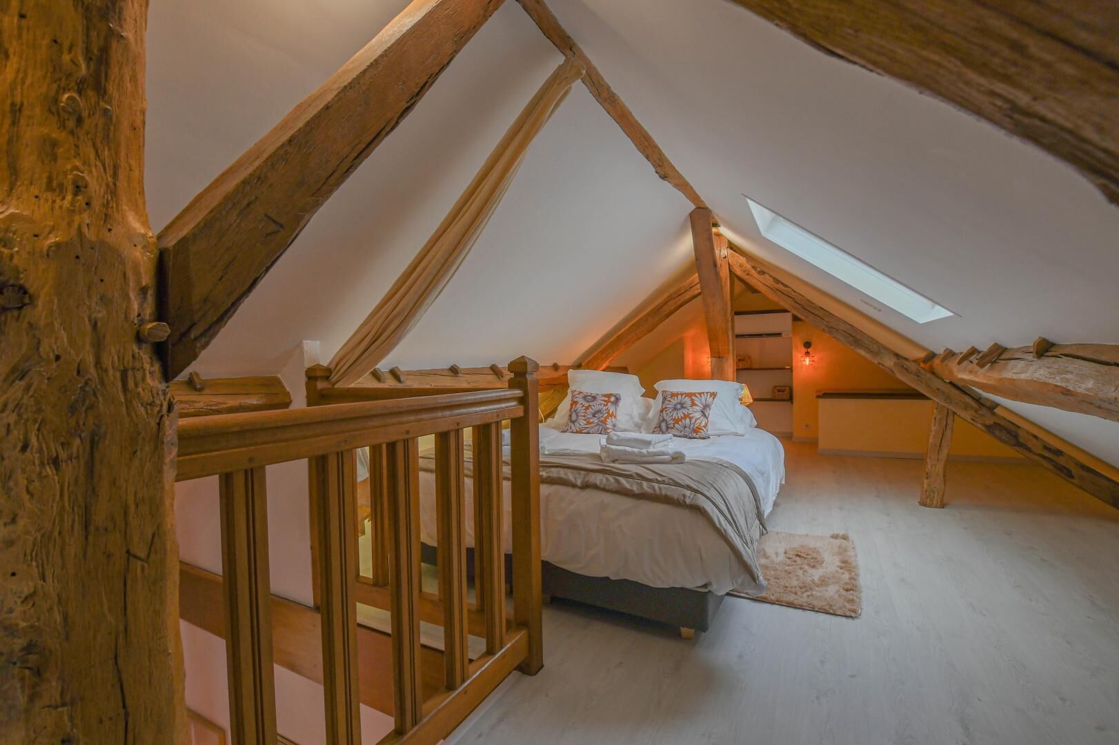 A cozy attic bedroom with a sloped ceiling, wooden beams, a bed with floral pillows, and a skylight.
