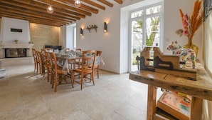 A dining area with a wooden table set for six, surrounded by chairs. There is a fireplace with a stone wall behind it, and a large window providing natural light. - Le Clos Embelli - ROUVRES LES VIGNES (ROUVRES LES VIGNES)