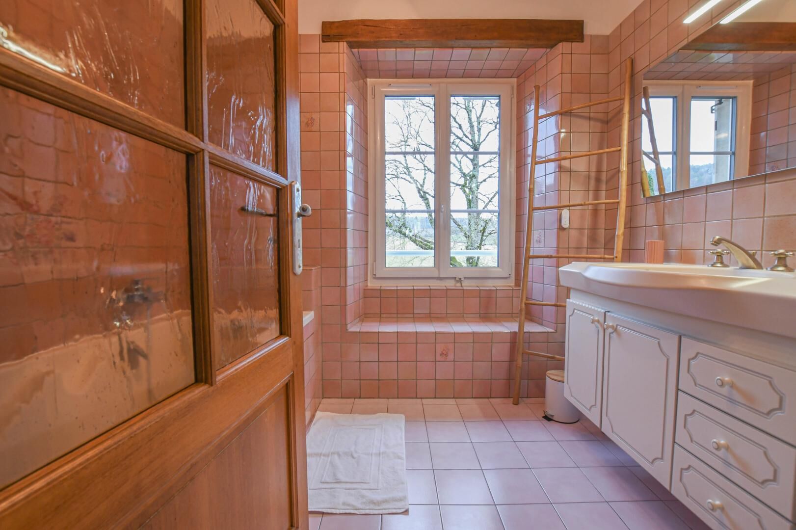 Un baño con puerta de madera, ventana con vistas, lavabo y espejo.