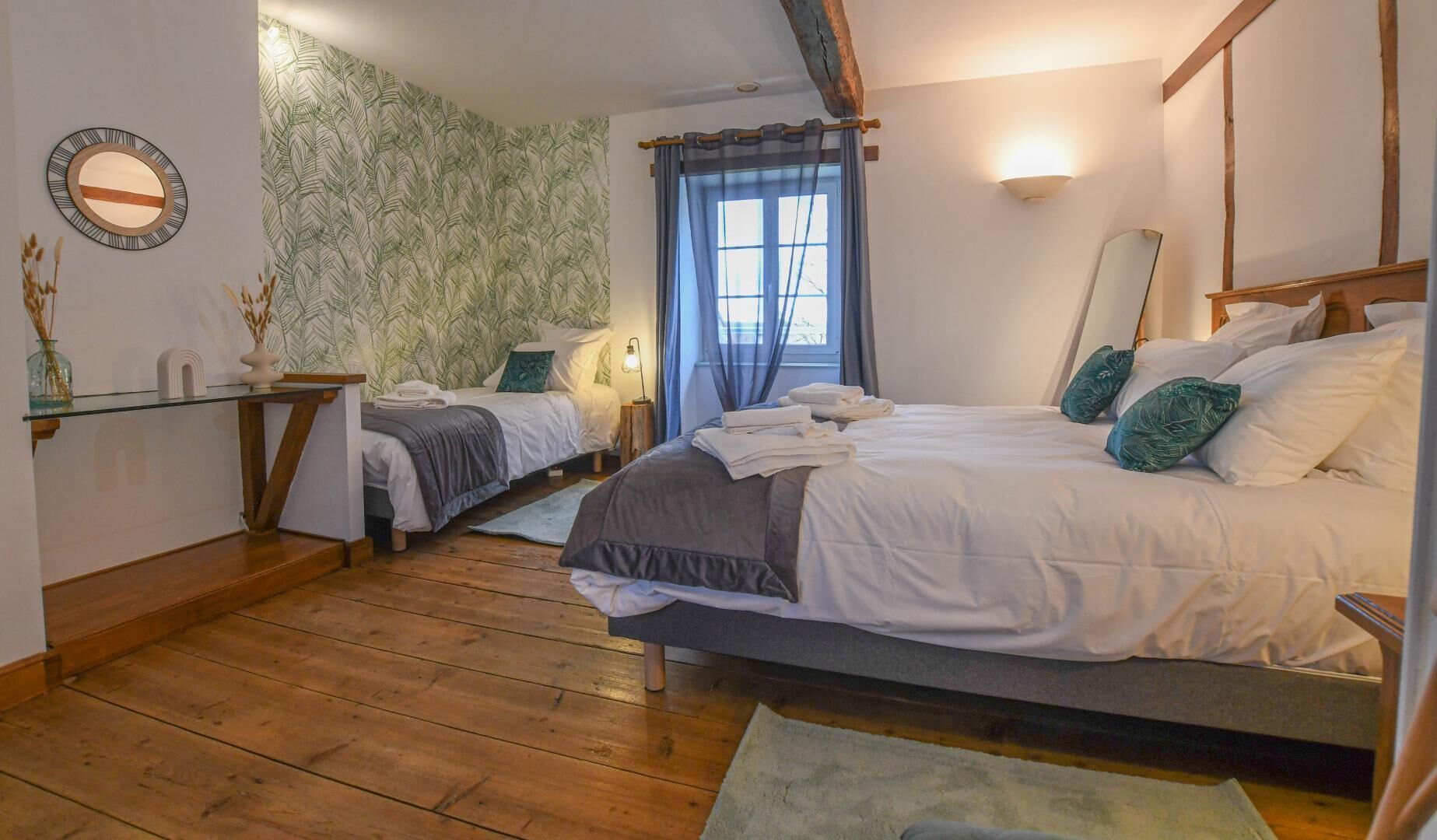 Camera da letto con due letti, pavimento in legno, parete con motivo a foglia, comodino con lampada e finestra con tende.