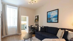Ett modernt vardagsrum med en mörk soffa, ett litet bord och en stol. Det finns ett fönster med gardiner, en tavla på väggen och ett element.