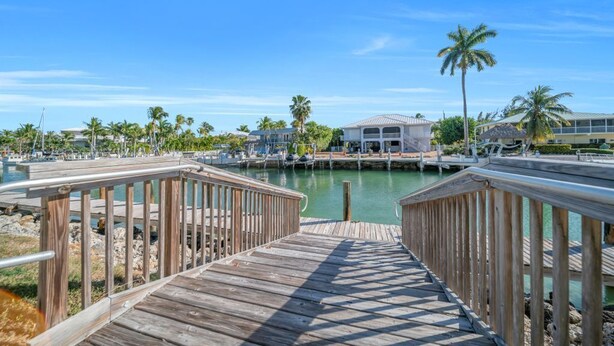 A wooden boardwalk leading to a marina with boats, palm trees, and a building with a white roof. - 7026 Waterfront Duck Key Villa - Sleeps 6 (Marathon)