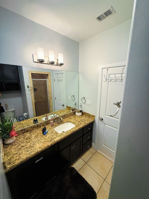 Une salle de bain avec un lavabo sur un plan de travail en granit, des meubles sombres, un miroir et un espace douche.
