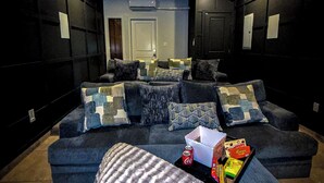 Un salón con decoración oscura, un sofá seccional, una mesa de centro con aperitivos y una pantalla de proyector.