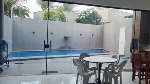 Ein moderner Poolbereich im Freien mit Glaswand, einem Esstisch mit Stühlen und einem Holzsitzset.