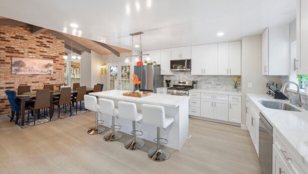 A modern kitchen with white cabinetry, a central island with bar stools, and a brick accent wall. - Monthly 4BD 2.5BR Home w Gorgeous View, Heated Pool, Billiards, 65' tv, Games + (Vallejo)