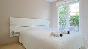 A bedroom with a large bed, a white headboard, a bedside table with a lamp, and a view of greenery through a window. - Le Maria - 2 bedroom - 2 Bath - Terrace (Cannes)