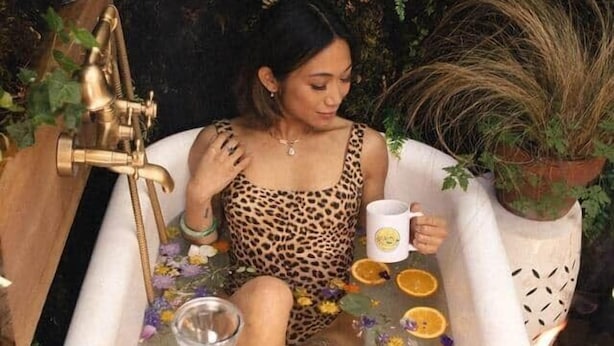 A person sitting in a bathtub filled with flowers and fruits, surrounded by plants and a small table with a candle and a bottle. - Chic 1967 Aristocrat Camper | Glamping (Tub) (Burnt Ranch)