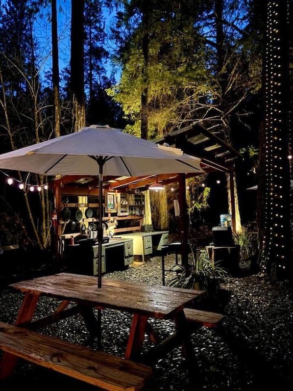 An outdoor dining area with a wooden table and benches, surrounded by trees and a covered seating area with string lights.