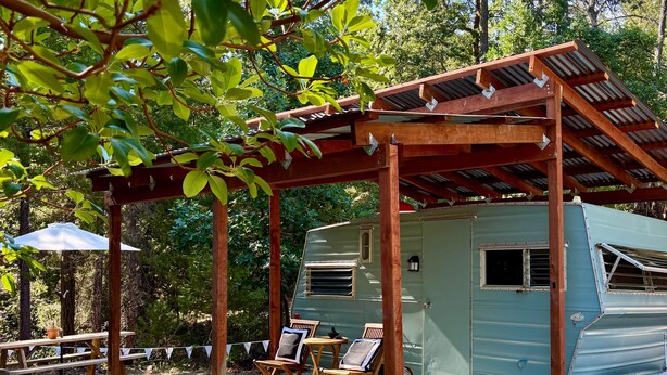 A rustic outdoor setting with a wooden pergola, a vintage camper, and a picnic table with chairs. - Chic 1967 Aristocrat Camper | Glamping (Tub) (Burnt Ranch)