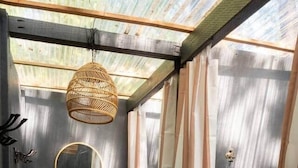 A small, enclosed space with a metal roof, a hanging woven light fixture, a mirror, striped curtains, a potted plant, and a basket labeled "TOWELS". - Chic 1967 Aristocrat Camper | Glamping (Tub) (Burnt Ranch)