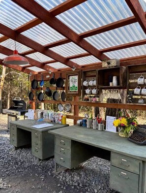 A rustic outdoor kitchen with a workbench, cooking utensils, and a grill.