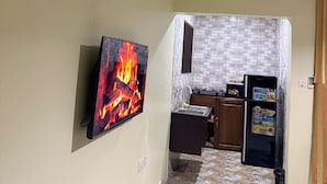 A room with a mounted air conditioner, a flat-screen TV displaying a fire, and a kitchen area with a tiled backsplash and cabinets.