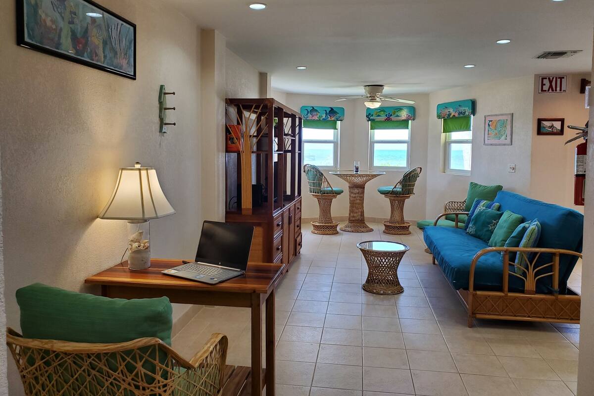 A room with a laptop on a wooden table, a blue sofa, wicker chairs, and a small round table.