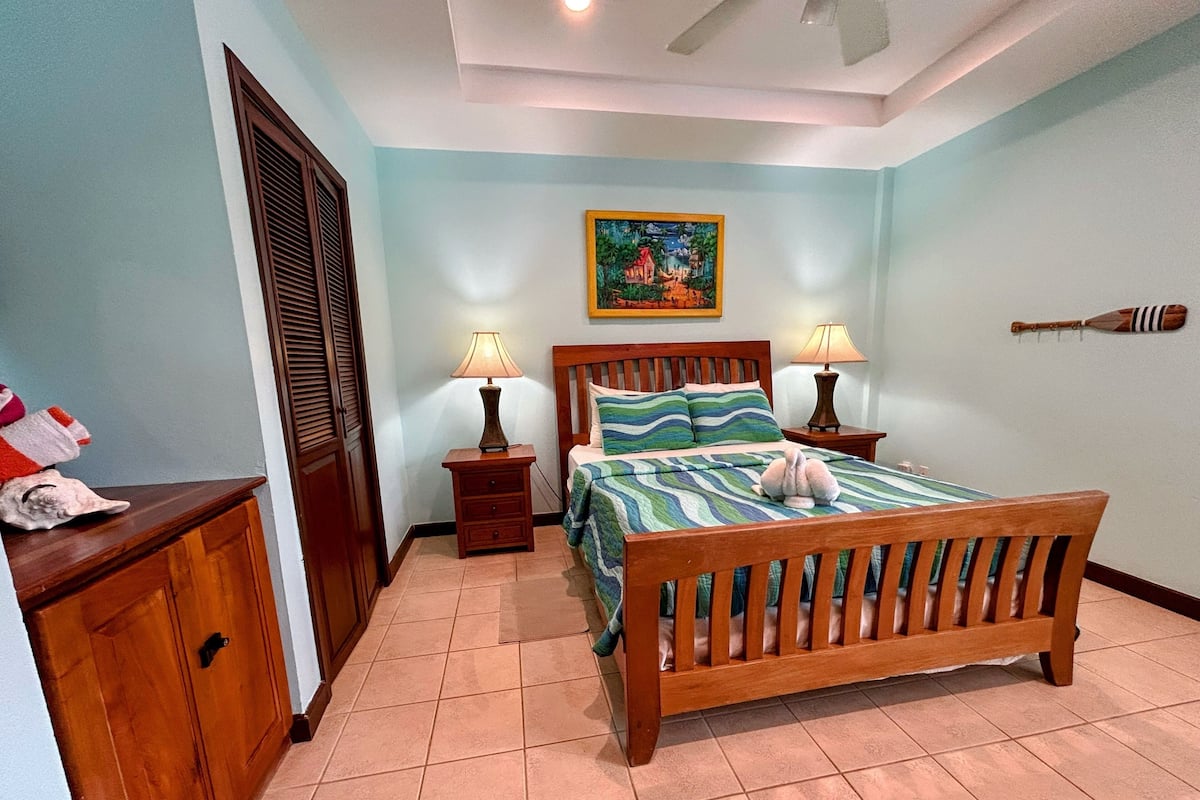 A bedroom with a wooden bed, two bedside tables with lamps, a wooden wardrobe, a painting on the wall, and a decorative wall hanging.
