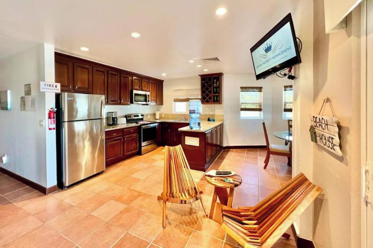 A modern kitchen with stainless steel appliances, wooden cabinets, and a flat-screen TV mounted on the wall.