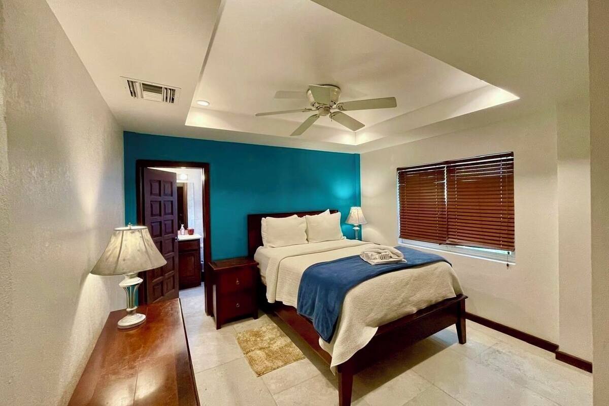 A bedroom with a bed, nightstands, a lamp, a ceiling fan, and a window with blinds.