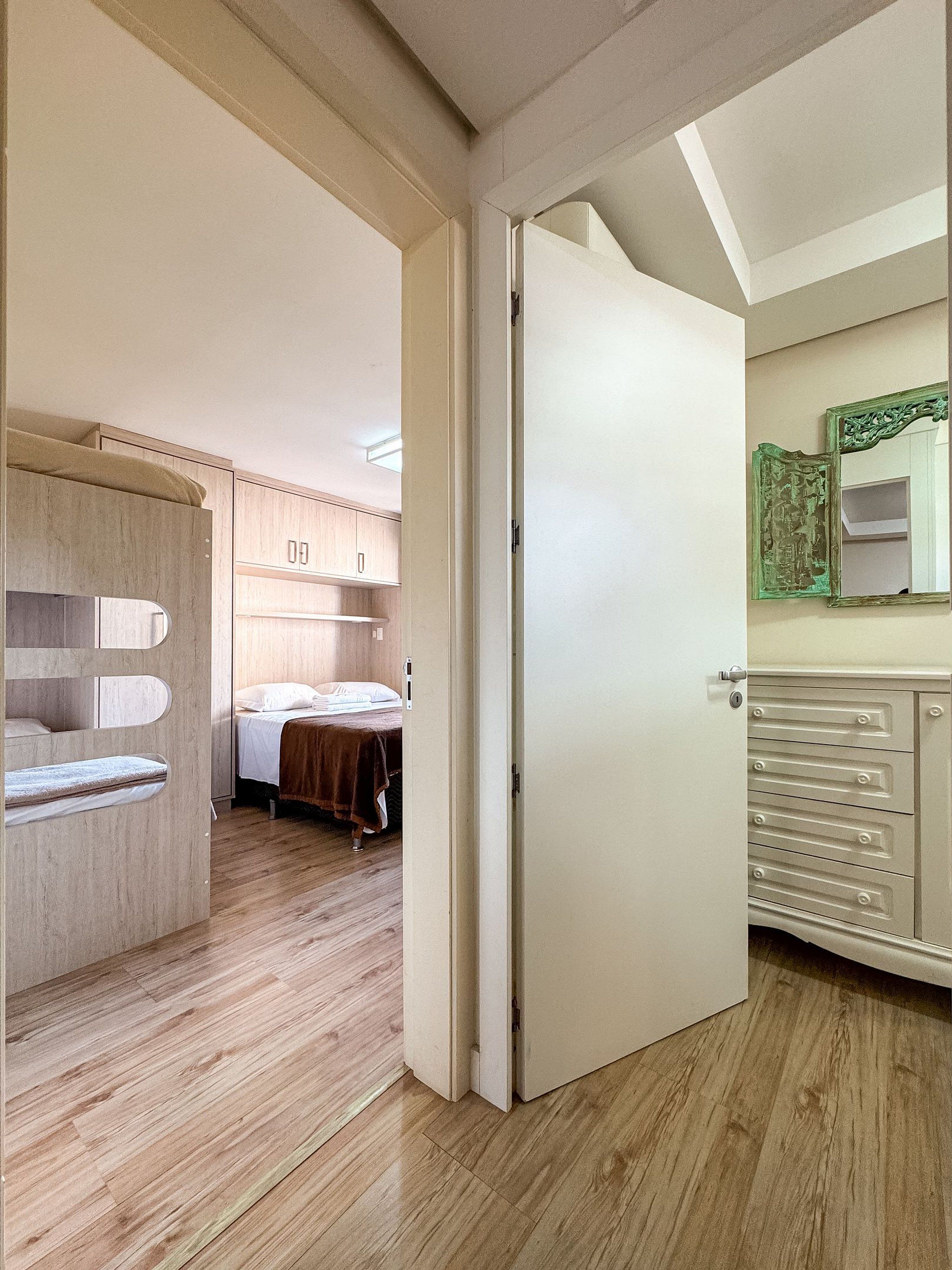 A hotel room with a wooden floor, a bed with a brown blanket, a wardrobe with multiple drawers, and a mirror with an ornate frame.
