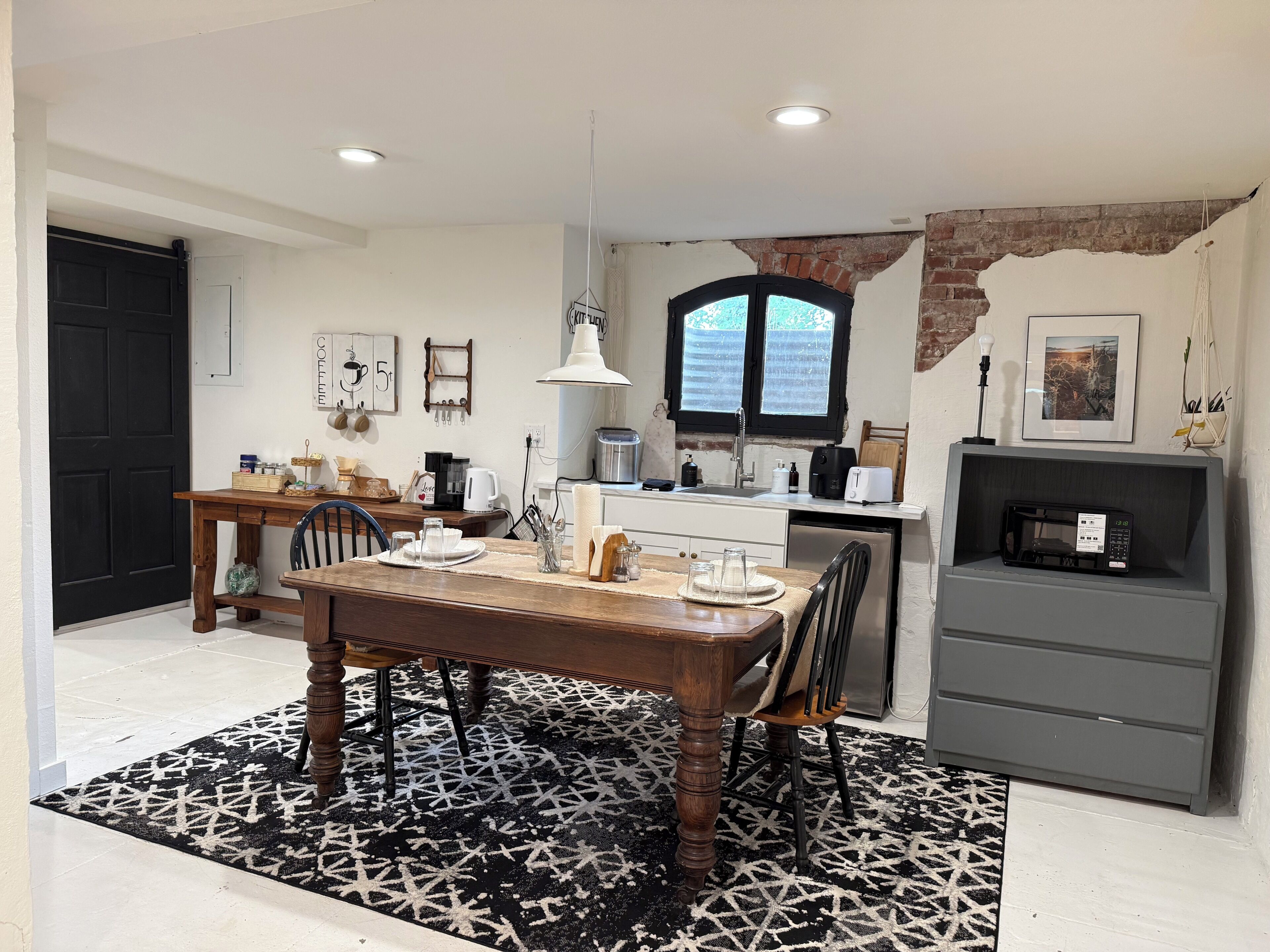 A modern kitchen with a dining area, featuring a wooden table, chairs, a black and white patterned rug, and a brick wall.