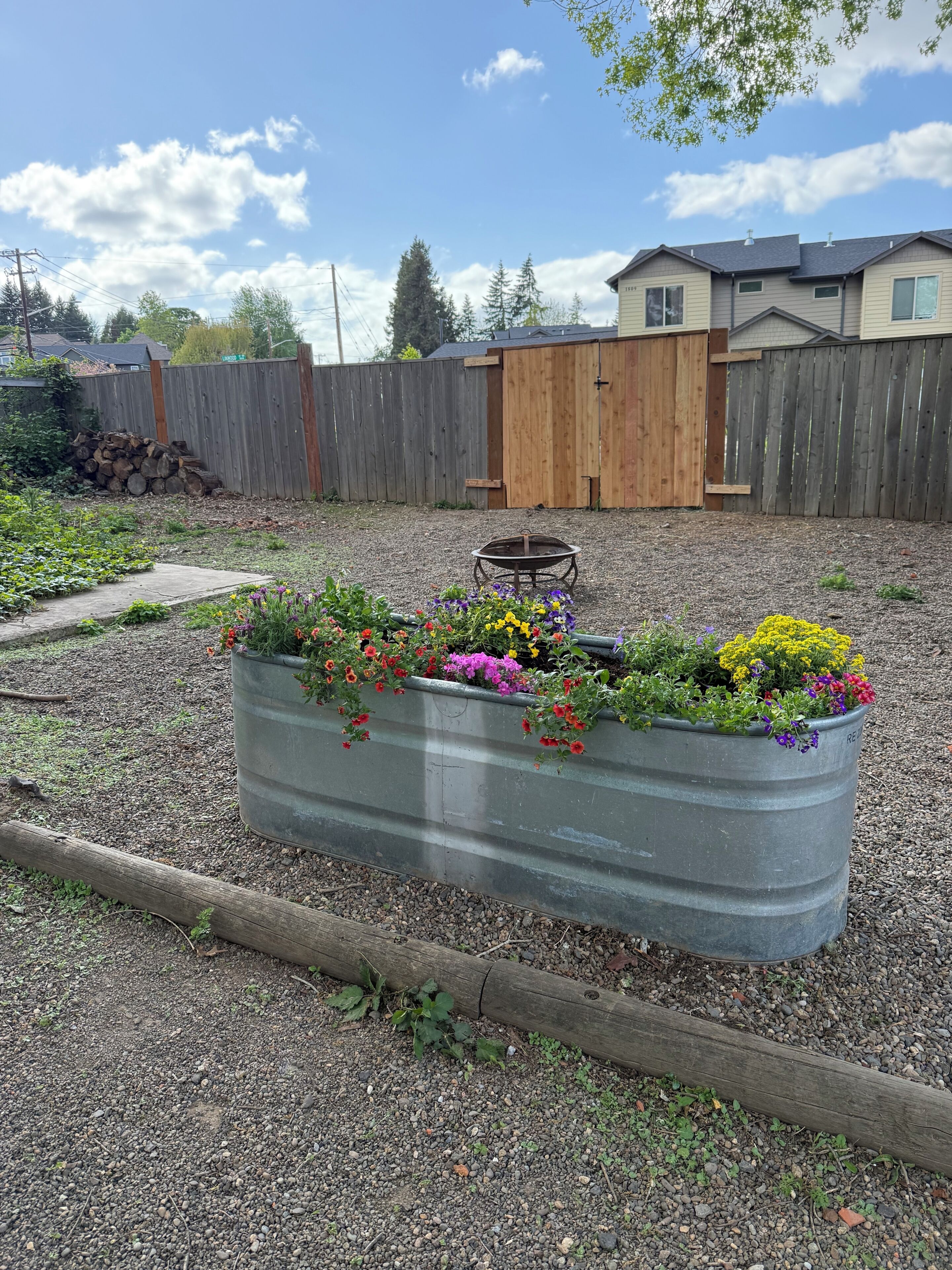 Een tuin met een metalen plantenbak vol kleurrijke bloemen, een houten omheining en een houten poort op de achtergrond.