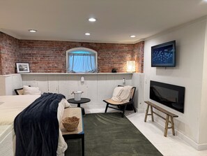 A hotel room with a brick wall, a bed, a chair, a small table, a TV, and a window with a curtain. - Farmhouse Suite in Willamette Valley (Salem)