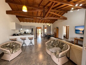 A spacious living area with a high ceiling and exposed wooden beams, a kitchen with modern appliances, and a cozy seating area with wicker furniture. - Casa 2 Quartos - Centro - Xangri-Lá (Xangri-La)