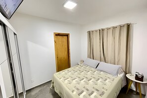 A bedroom with a bed, a wooden door, a window with curtains, a small table with a lamp, and a TV mounted on the wall.