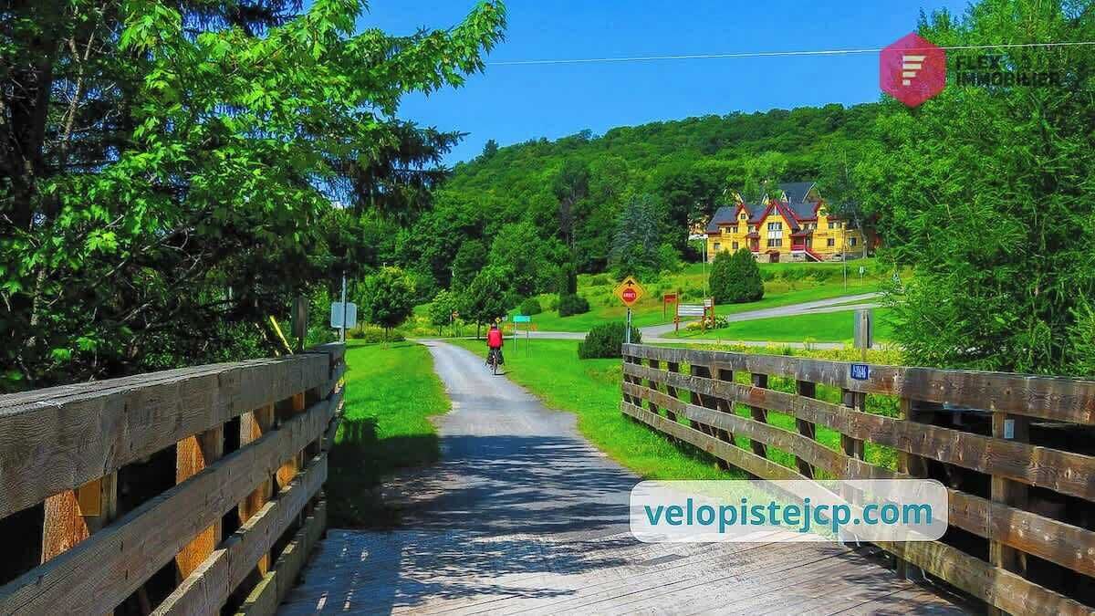 A wooden bridge over a narrow path with a cyclist in the distance, surrounded by lush greenery and a large house in the background.
