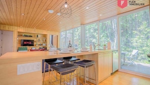 A modern kitchen with wooden cabinetry, a central island with bar stools, a built-in TV, and large windows offering a view of the outdoors.