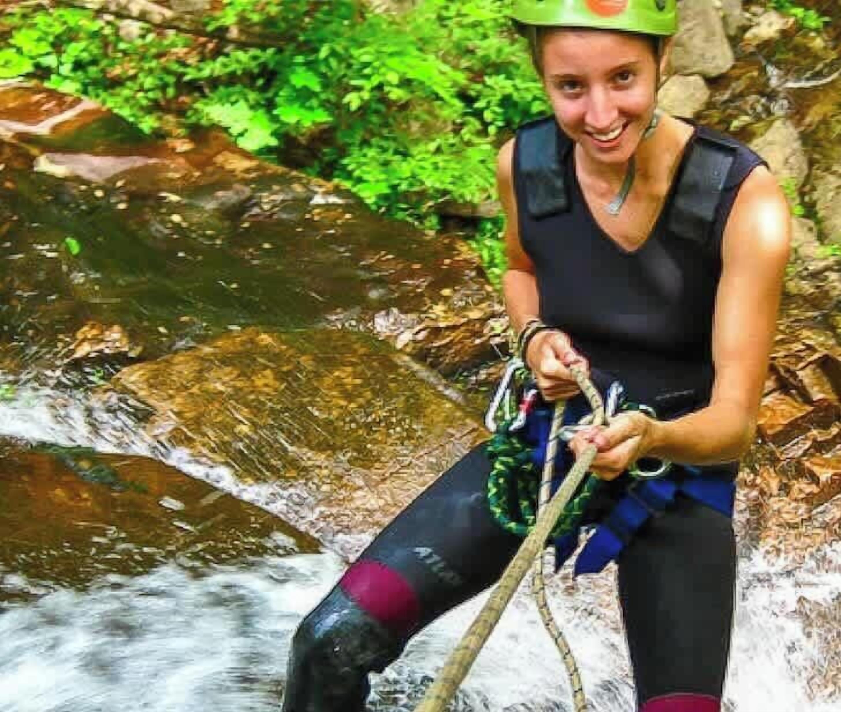A person in a black sleeveless top and waterproof pants, holding a rope, standing by a flowing stream in a natural setting.