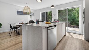 Een moderne keuken met een centraal eiland, grote ramen en een eetgedeelte met stoelen.