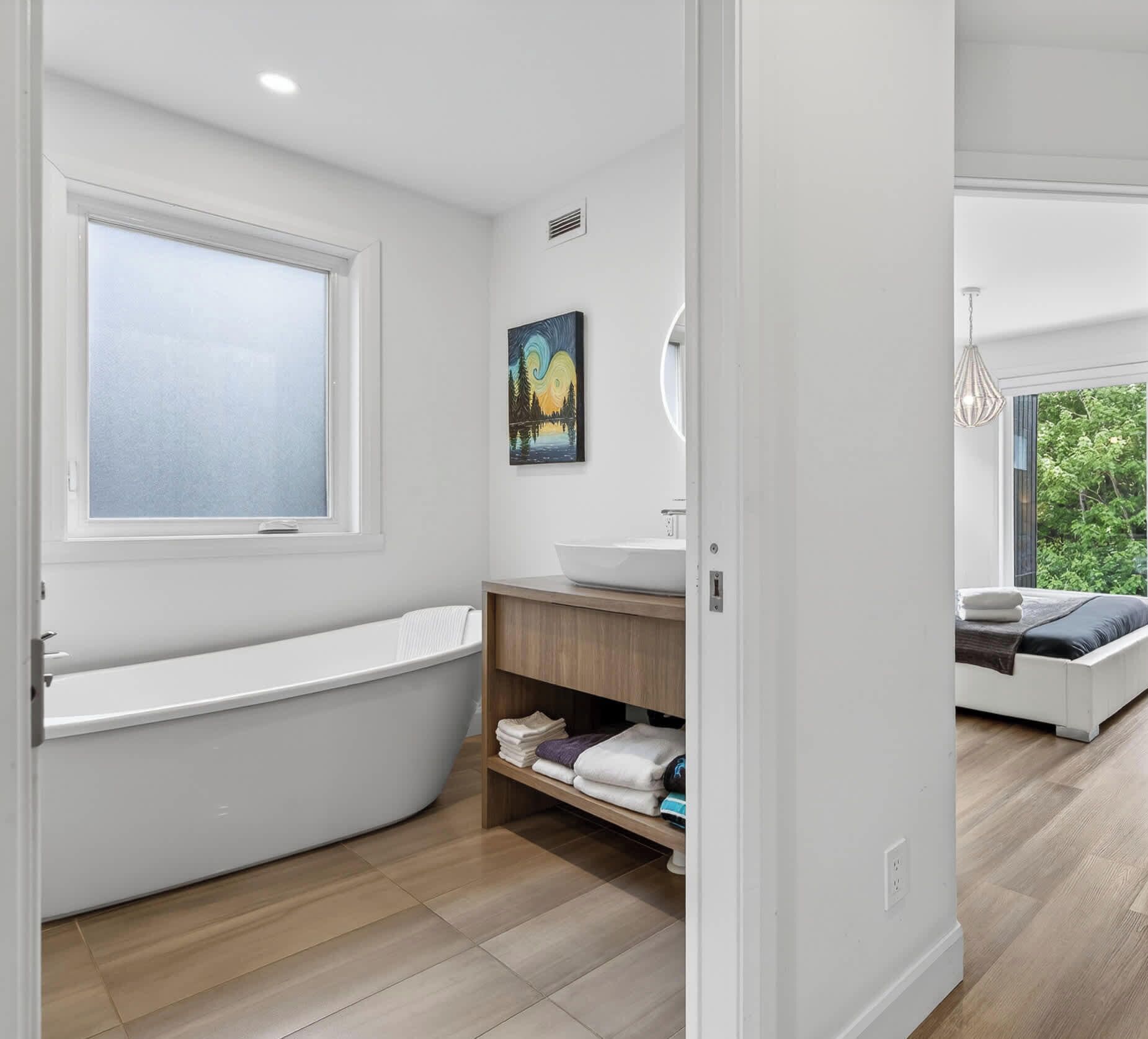 Une salle de bain moderne avec une baignoire, une étagère en bois avec des serviettes, un tableau et une vue sur une chambre à coucher avec un lit et une fenêtre.
