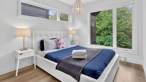 Een slaapkamer met een groot raam, een bed met een blauw-grijze spreideken, twee nachtkastjes met lampen, en een hangende lamp.
