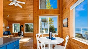 Une cabane en bois comprenant un coin repas, des chaises blanches, une table, un ventilateur de plafond et de grandes fenêtres donnant sur l’océan.