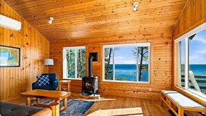 Un intérieur de chalet chaleureux avec des murs en bois, une cheminée, un canapé, une table et de grandes fenêtres offrant une vue sur l’océan.