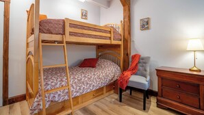 Una cama litera con una cama inferior, una silla, una lámpara y un aparador en una habitación con piso de madera y paredes blancas.