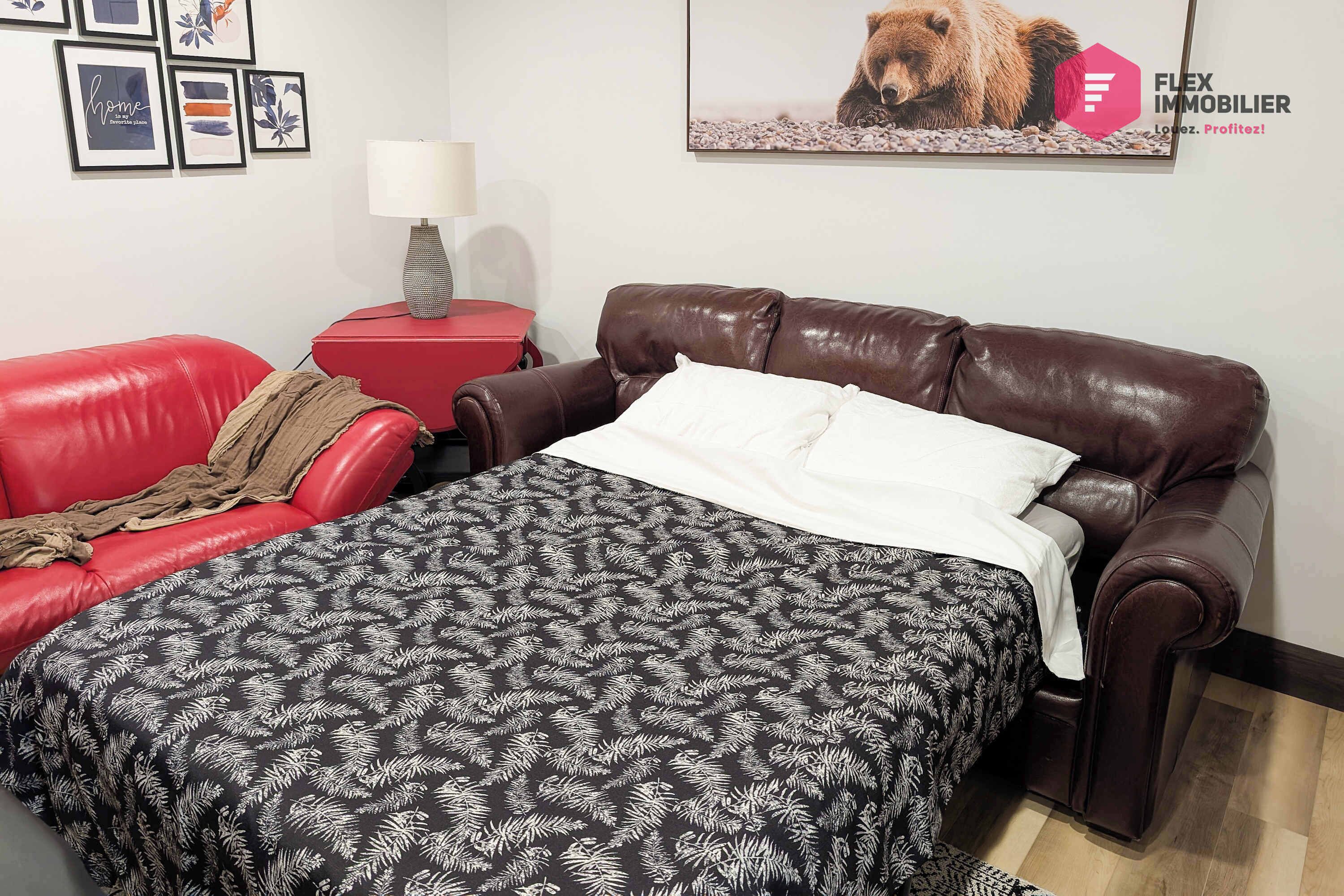 A bedroom with a brown leather sofa, a red side table, a lamp, and a bed with a patterned bedspread.
