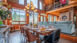 Comedor con mesa de madera preparada para cuatro personas, rodeada de sillas y vistas a árboles a través de amplios ventanales.