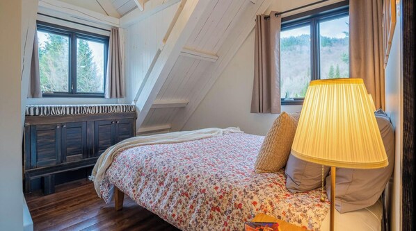 A bedroom with a sloped ceiling, a bed with floral bedding, a bench with a striped cushion, and a lamp on the bedside table. - Stoneham St-Évasion, Waterfront Sauna spa for 9 (Québec)