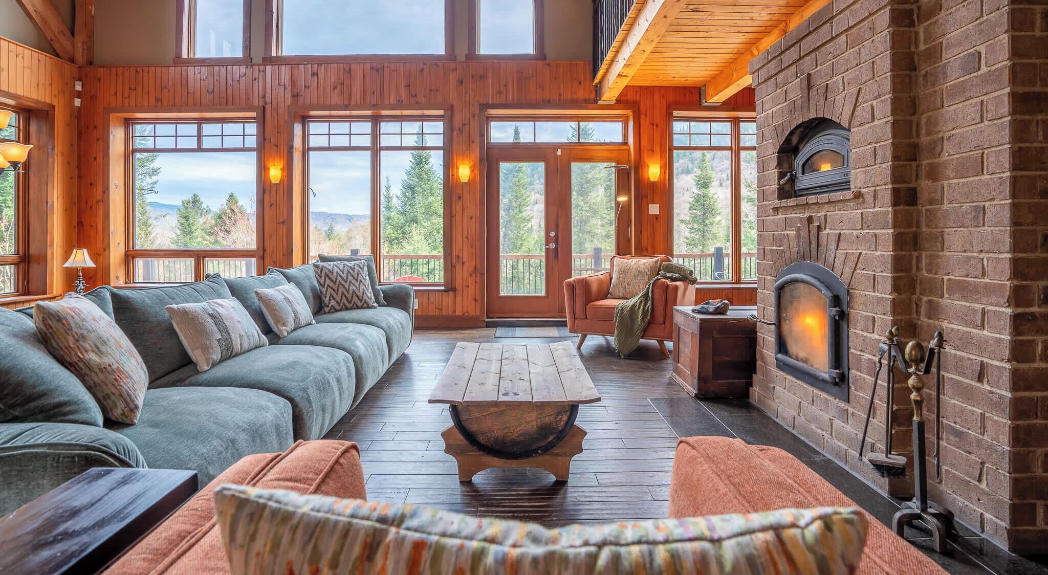 A cozy living room with a fireplace, wooden flooring, and large windows offering a view of the outdoors.