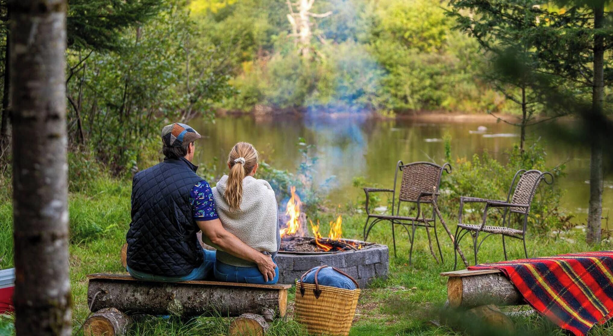 Ett par sitter vid ett lägerbål i ett skogsområde, med en sjö i bakgrunden.