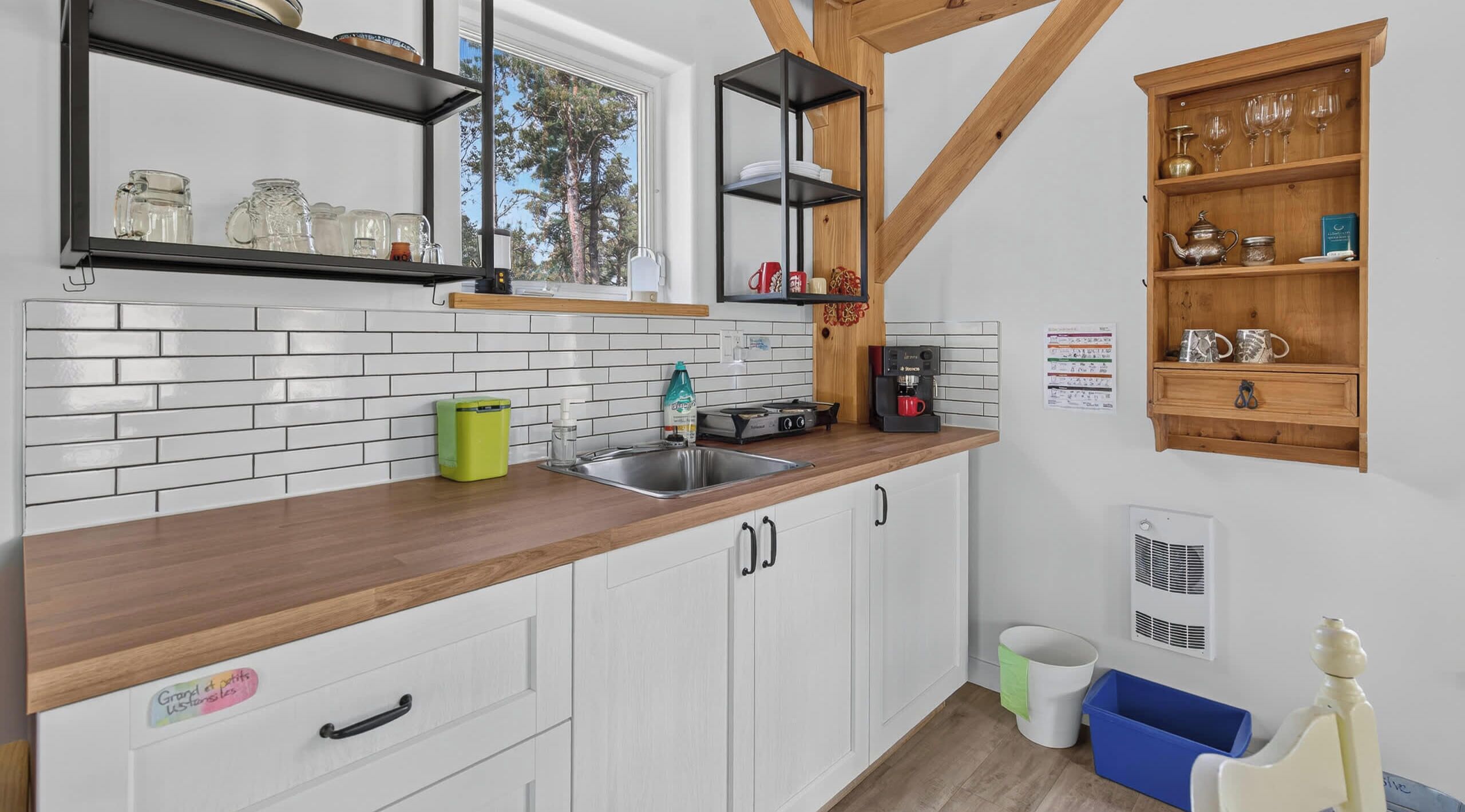 Una cocina con armarios blancos, encimera de madera, cafetera y ventana con vistas a árboles.