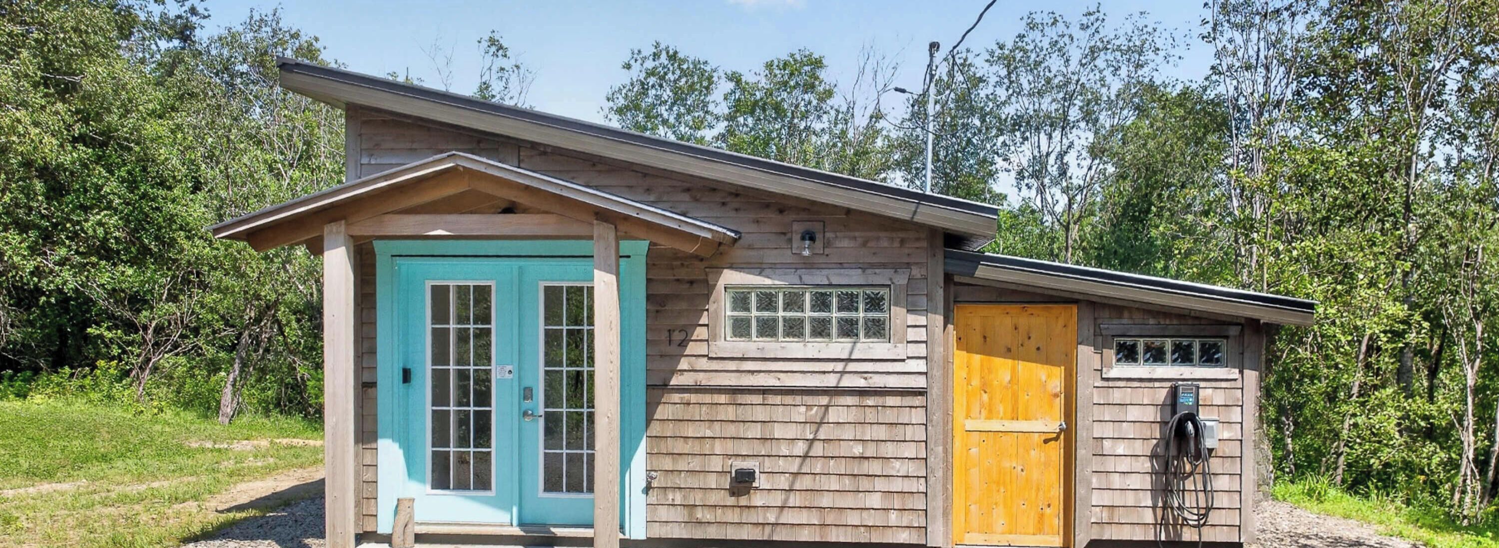 Een klein houten huisje met een turquoise deur en een gele deur, omringd door bomen.
