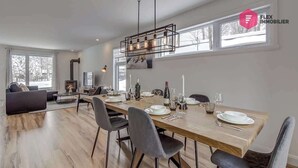 A spacious living room with a large wooden dining table set for six, a fireplace, and a modern chandelier. - The Urban Flower in Portneuf - SPA, Sauna, Pool (Pont-Rouge)