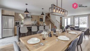 A modern kitchen with a dining area set for a meal, featuring stainless steel appliances, wooden cabinets, and a large dining table with chairs. - The Urban Flower in Portneuf - SPA, Sauna, Pool (Pont-Rouge)